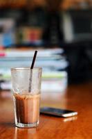 A glass of iced chocolate on wooden table with blurred smartphone and magazines  background. photo