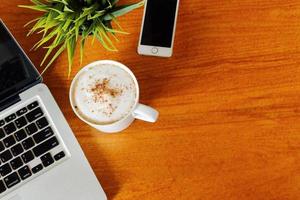 computadora portátil, teléfono inteligente y una taza de café con leche caliente en la vista superior de la mesa de madera. foto