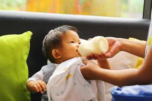 Asian baby sitting on sofa and drinking milk from baby bottle feeding by mother. photo