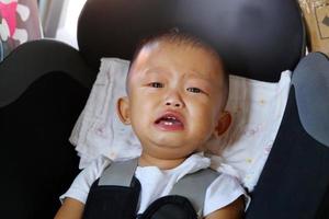 Little baby boy crying while fastened in safety car seat. Asian child traveling by car. photo