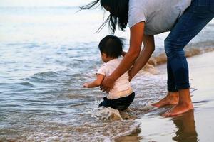 bebé asiático juega ola de mar con madre en la playa. niño juega con sus padres en la playa. foto