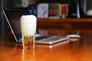 Glass of beer on wooden table with computer notebook in cafe. Beer in mug on table with laptop background in bar. photo