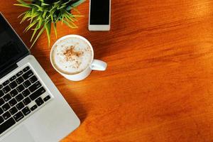 café con leche caliente en una taza en la vista de la mesa de madera desde arriba con una computadora portátil y un teléfono inteligente alrededor tienen espacio para copiar. foto