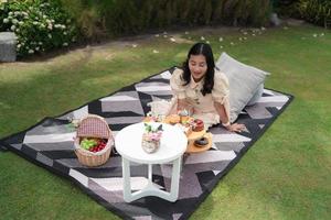 happy Asian woman excited with dessert during picnic in fresh green garden, picnic set with snack and pillow
