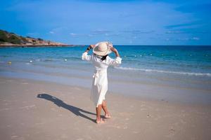 white dress woman in the back with wide brim hat walking to the sea from sand beach
