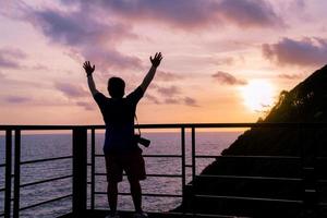 silhouette freedom Asian man photographer standing at terrace on top hill with cliff and sea view in evening with sunset twilight sky photo