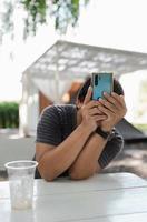 man in causal t-shirt holding mobile phone up to taking photo during sit on white wooden table after drinking coffee in camping garden, relax lifestyle