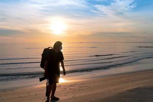 silueta hombre asiático mochilero con mascarilla protectora sostenga la cámara caminando en la playa con el cálido cielo del amanecer matutino en huahin foto