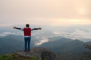 man with freedom arms open standing on rock ledge of mountain peak at Pha Tang view point 104 with river view and sea mist and sun rise in heavy fog sky. travel place in Chaing Rai, Thailand photo