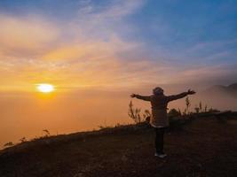 Asian woman with freedom arms open standing on mountain peak at Pha Tang view point 104 with river view and sea mist and sun rise in fog sky. travel place in Chaing Rai, Thailand photo