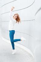 Beautiful girl with a smile in a white sweater, blue jeans and white sneakers jumping. Happy outdoor portrait on the background of modern metal wall. photo