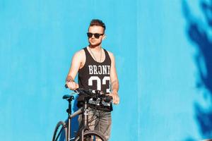 hombre hipster con gafas de sol con una bicicleta cerca de una pared azul brillante. foto