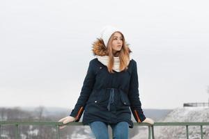 Cute girl in winter cap and jacket sitting on the railing. Outdoors in winter. photo