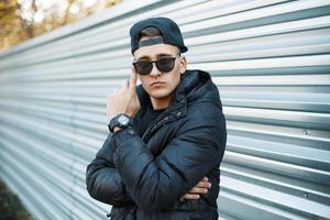 A young man in a stylish winter jacket and sunglasses on the background of a metal wall. photo