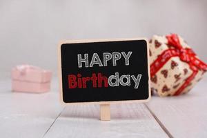 Black chalkboard with the Happy Birthday text on the table on the background with gifts. photo