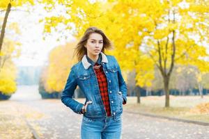 linda chica con una chaqueta de mezclilla y jeans en el fondo del follaje amarillo, un hermoso día de otoño foto