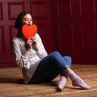 mujer de pelo corto con los ojos cerrados y los labios dando forma a un beso sentada en el suelo con las piernas cruzadas sosteniendo la forma del corazón frente a su barbilla foto