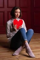 mujer de pelo corto mirando a la cámara sentada en el suelo con las piernas cruzadas sosteniendo la forma del corazón frente a ella foto