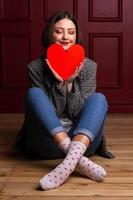 mujer sonriente de pelo corto con chaqueta sentada en el suelo con las piernas cruzadas sosteniendo forma de corazón frente a su barbilla foto