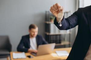 Smiling young modern man in a suit holds the keys to the bought apartment on the background of the realtor and office photo