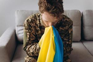 Portrait of emotional young Ukrainian patriot soldier in military uniform sitting on office on sofa holding hands and kissing yellow and blue flag. photo