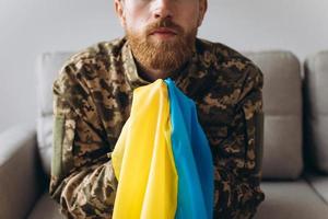 Portrait of an emotional young Ukrainian patriot soldier in military uniform sitting on the office on the couch holding a yellow and blue flag. photo