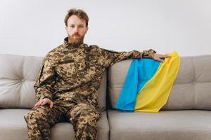 retrato de un emotivo joven soldado patriota ucraniano con uniforme militar sentado en la oficina en un sofá con una bandera amarilla y azul. foto