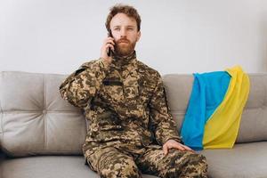 A Ukrainian soldier in military uniform is sitting on a sofa in the office and talking on the phone photo