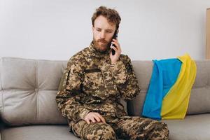 A Ukrainian soldier in military uniform is sitting on a sofa in the office and talking on the phone photo