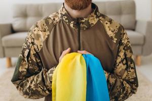 Portrait of an emotional young bearded Ukrainian patriot soldier in military uniform holding a flag standing on his knees photo