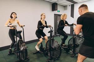 las mujeres jóvenes hacen ejercicio en bicicletas de aire en el gimnasio con entrenador motivador. foto