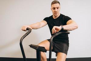 hombre fuerte mirando reloj y usando bicicleta de aire para hacer ejercicio cardiovascular en el gimnasio de entrenamiento cruzado. foto