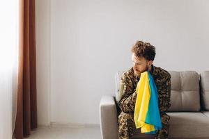 retrato de un emotivo joven soldado patriota ucraniano con uniforme militar sentado en la oficina en el sofá sosteniendo una bandera amarilla y azul.
