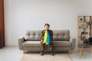 Portrait of an emotional young Ukrainian patriot soldier in military uniform sitting on the office on the couch holding a yellow and blue flag. photo