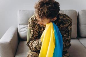 retrato de un emotivo joven soldado patriota ucraniano con uniforme militar sentado en la oficina en el sofá sosteniendo una bandera amarilla y azul. foto