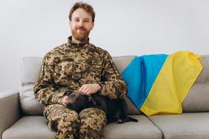 A Ukrainian soldier in military uniform is sitting on a sofa with his faithful friend, an Amstaff dog, on the background yellow and blue flag. photo