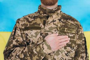 soldado patriota ucraniano con uniforme militar sostiene una mano en un corazón contra el fondo de una bandera amarilla y azul foto