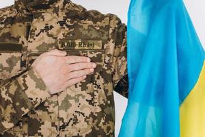 Ukrainian patriot soldier in military uniform holds a hand on a heart with a yellow and blue flag on a white background photo