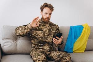 A Ukrainian soldier in military uniform is sitting on a sofa in the office and talking on the phone photo