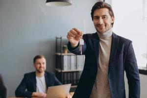 Smiling young modern man in a suit holds the keys to the bought apartment on the background of the realtor and office photo