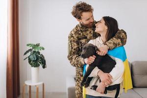 una pareja ucraniana, un soldado con uniforme militar y una niña envuelta en una bandera ucraniana sostienen un perro en sus brazos, felices juntos. foto