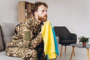 retrato de un emotivo joven soldado patriota ucraniano con uniforme militar sentado en la oficina en el sofá sosteniendo una bandera amarilla y azul. foto