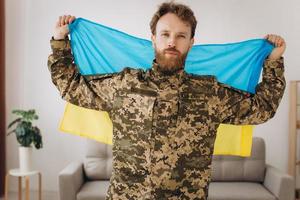 Portrait of an emotional young bearded Ukrainian patriot soldier in military uniform holding a flag in the office photo