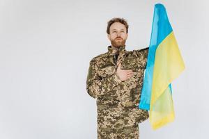 Ukrainian patriot soldier in military uniform holds a hand on a heart with a yellow and blue flag on a white background photo