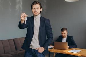 Smiling young modern man in a suit holds the keys to the bought apartment on the background of the realtor and office photo