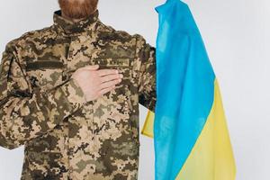 Ukrainian patriot soldier in military uniform holds a hand on a heart with a yellow and blue flag on a white background photo