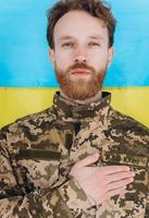 Ukrainian patriot soldier in military uniform holds a hand on a heart against the background of a yellow and blue flag photo