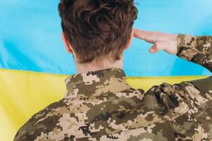 Ukrainian patriot soldier in uniform holds his hand to his head against the background of a yellow and blue flag photo