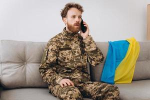 A Ukrainian soldier in military uniform is sitting on a sofa in the office and talking on the phone photo