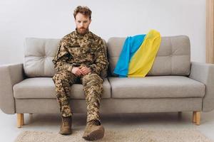 Portrait of an emotional young Ukrainian patriot soldier in military uniform sitting on the office on a sofa with a yellow and blue flag. photo